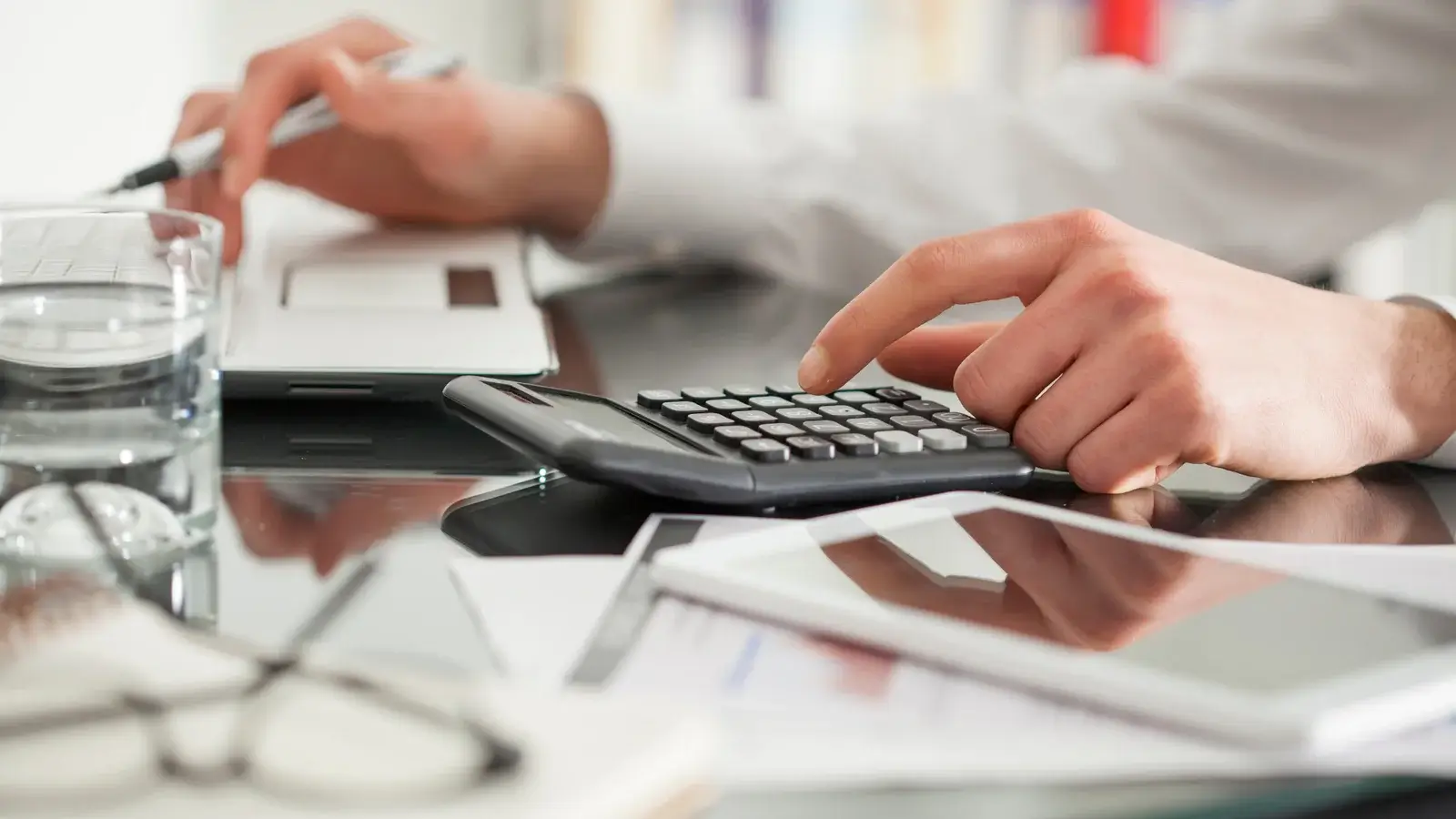 Uma pessoa fazendo cálculos e organizando gastos com uma calculadora, computador para anotar e contas em cima da mesa.